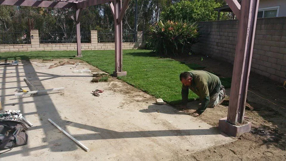 Worker grading compacted soil base for patio installation under wooden pergola